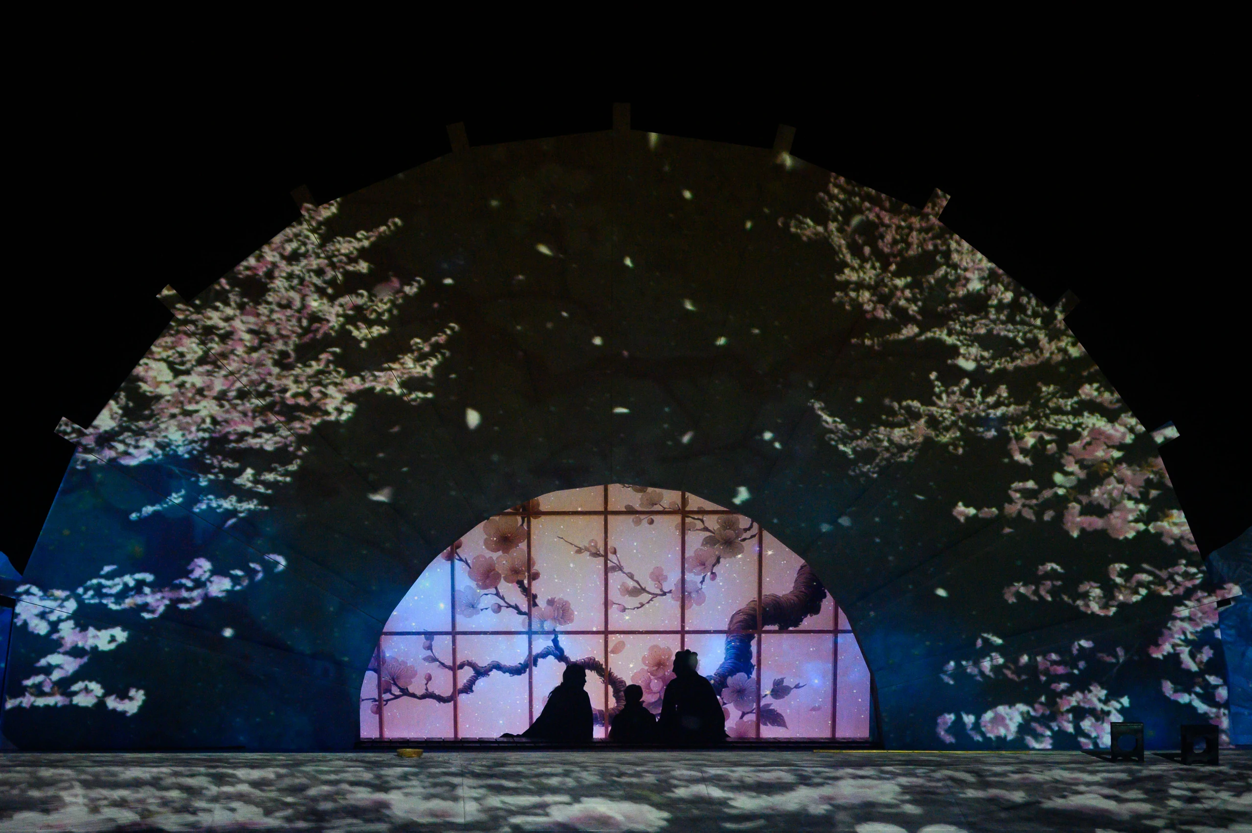 Scena di Madama Butterfly al Festival Puccini, con tre figure in silhouette e proiezioni di fiori di ciliegio